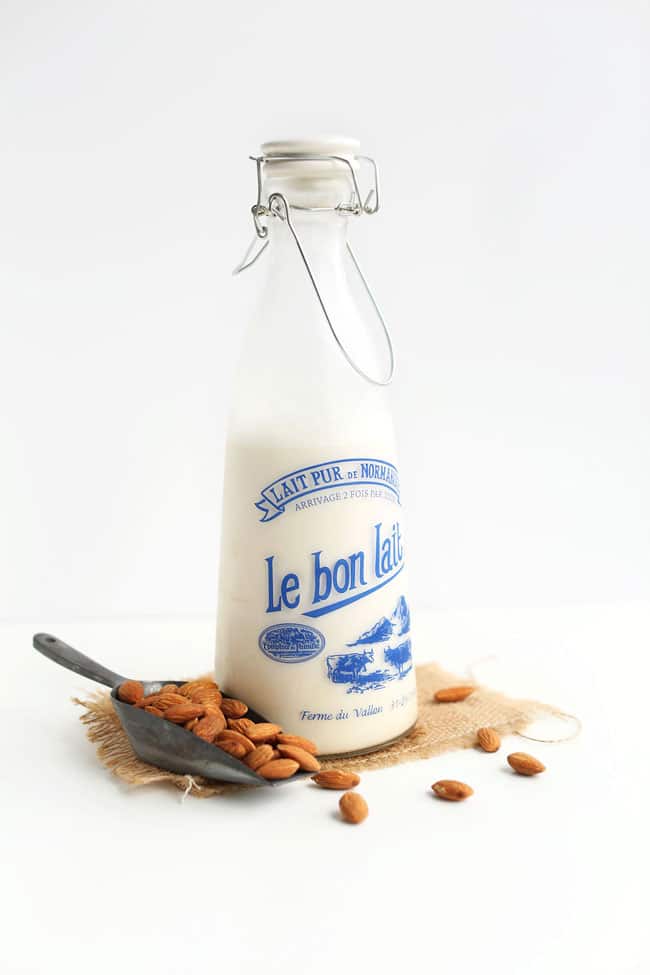 A glass bottle of almond milk sitting on a white background with a few almonds strewn about.