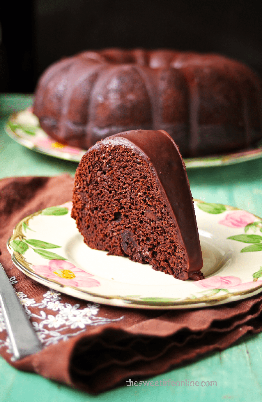 A slice of chocolate bundt cake sitting on a flowered plate with a cloth napkin under it and the rest of the cake in the background.
