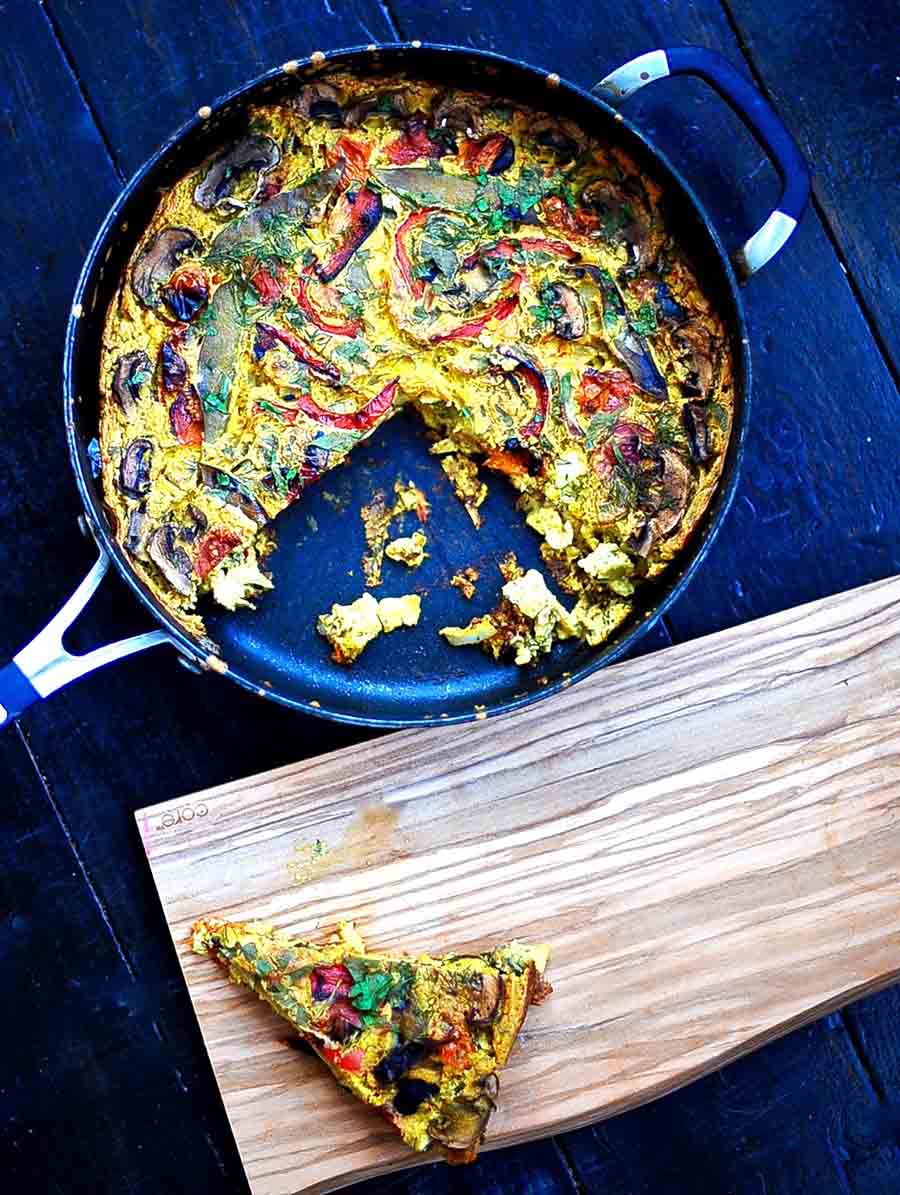 An overhead view of a vegetable frittata in a pan with a slice taken out and sitting next to it on a wooden board.